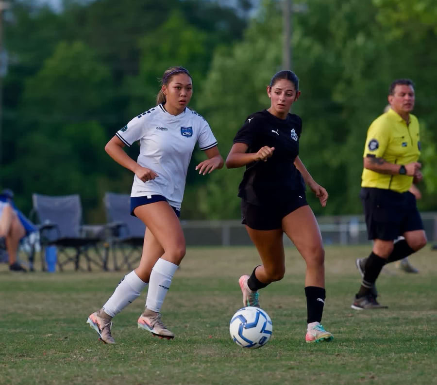 Female soccer player in orange jersey argues with referee on the outdoor field during a sunny day match.