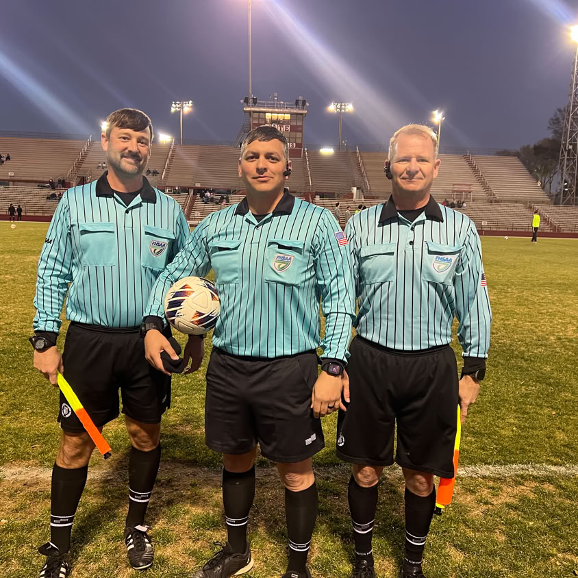 Three referees in green uniforms on a football field during a match.